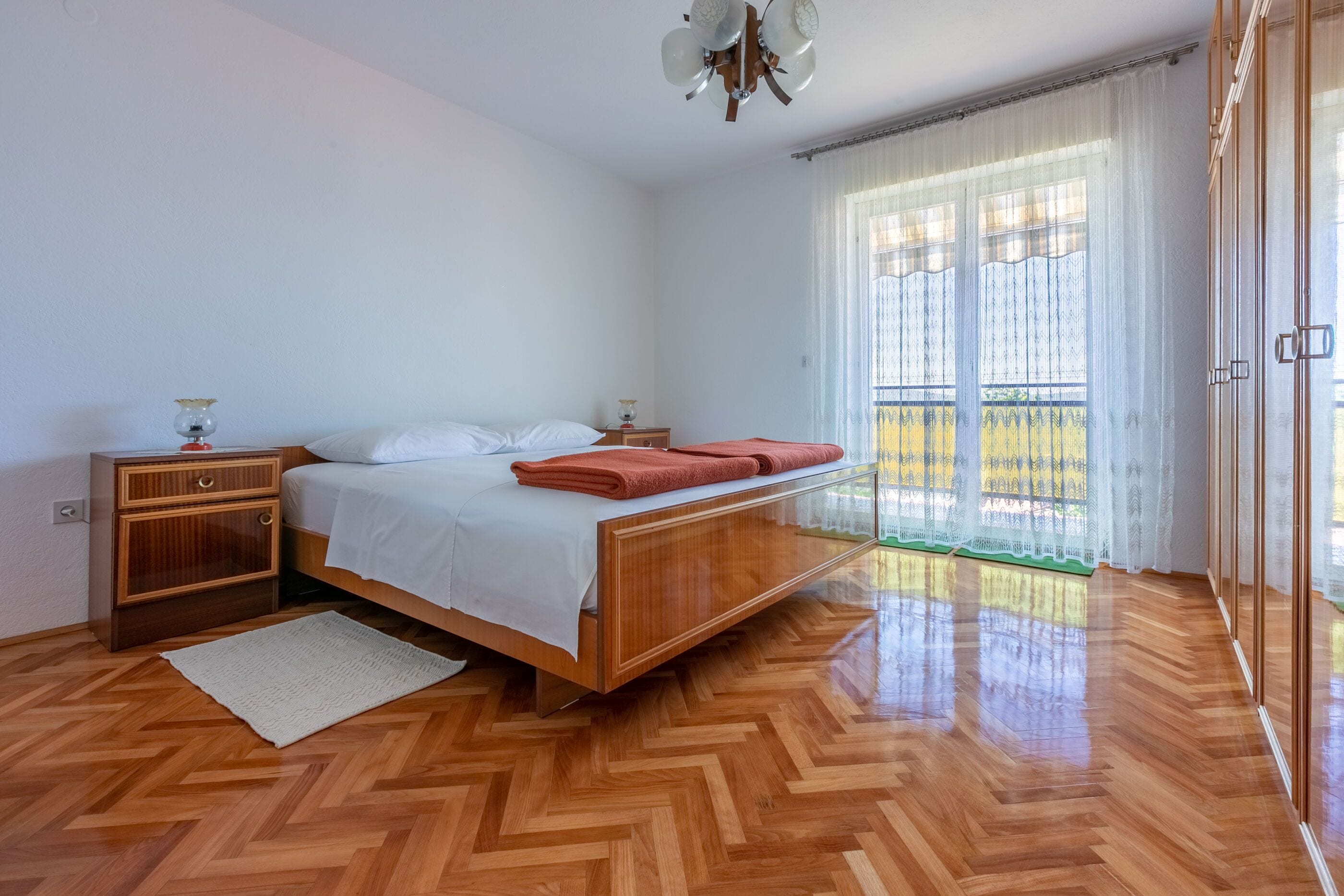 A bedroom with a wooden bed, a nightstand, a lamp, and a view of a balcony through sheer curtains.