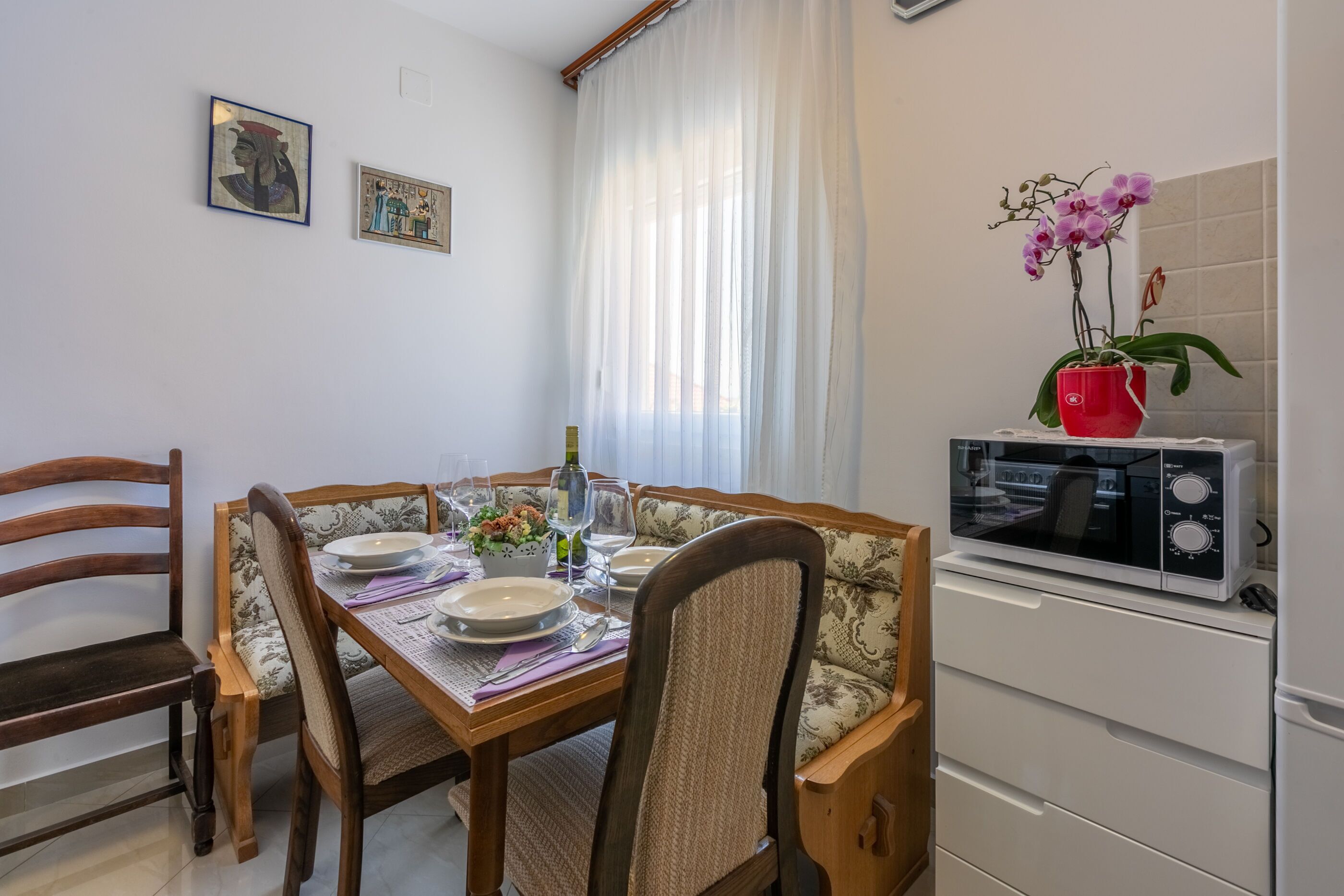 A dining area with a wooden table set for four, chairs, a vase with flowers, and a microwave on a cabinet.