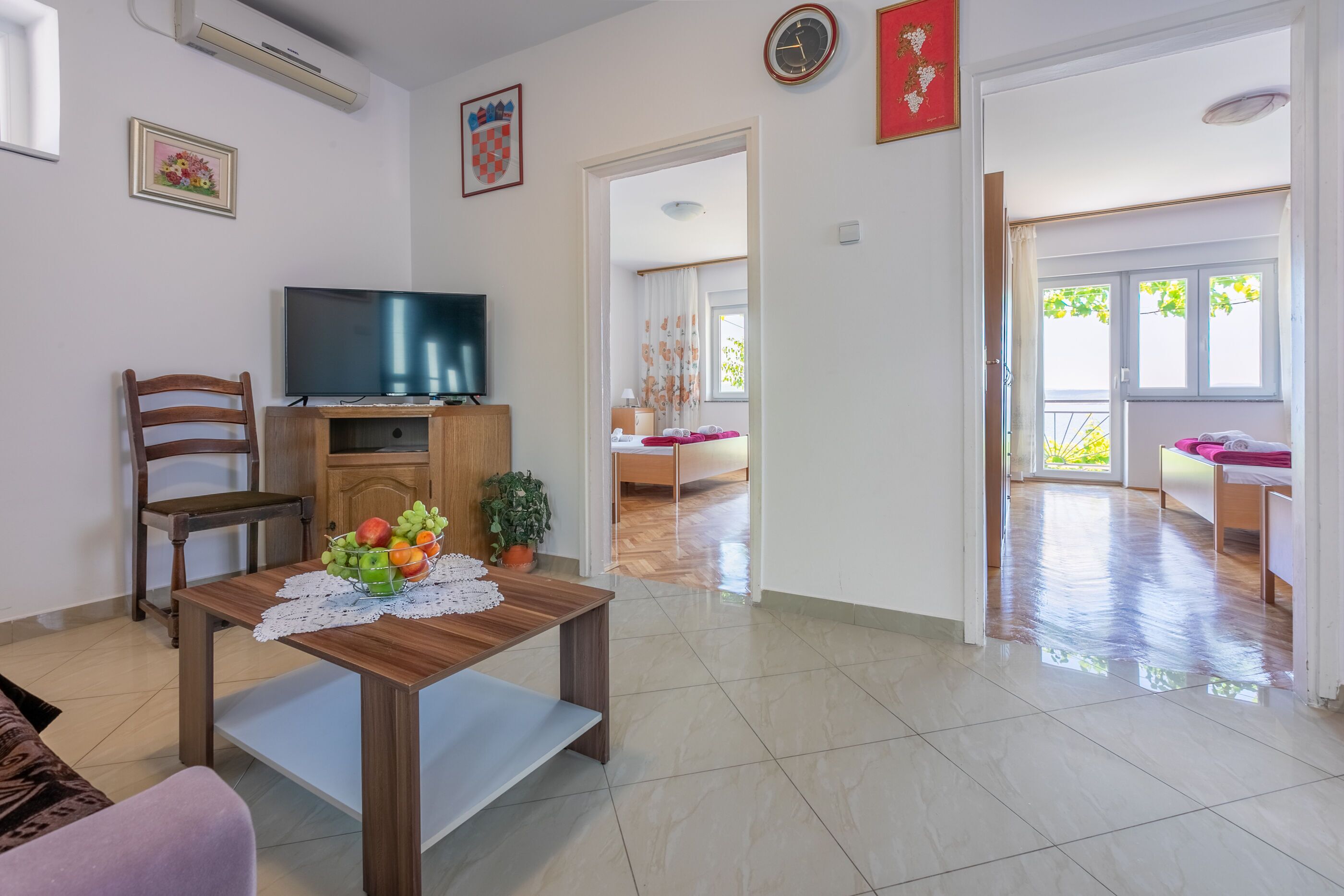 Una sala de estar con una mesa de madera, un televisor sobre un mueble bajo, una silla y una planta en maceta.