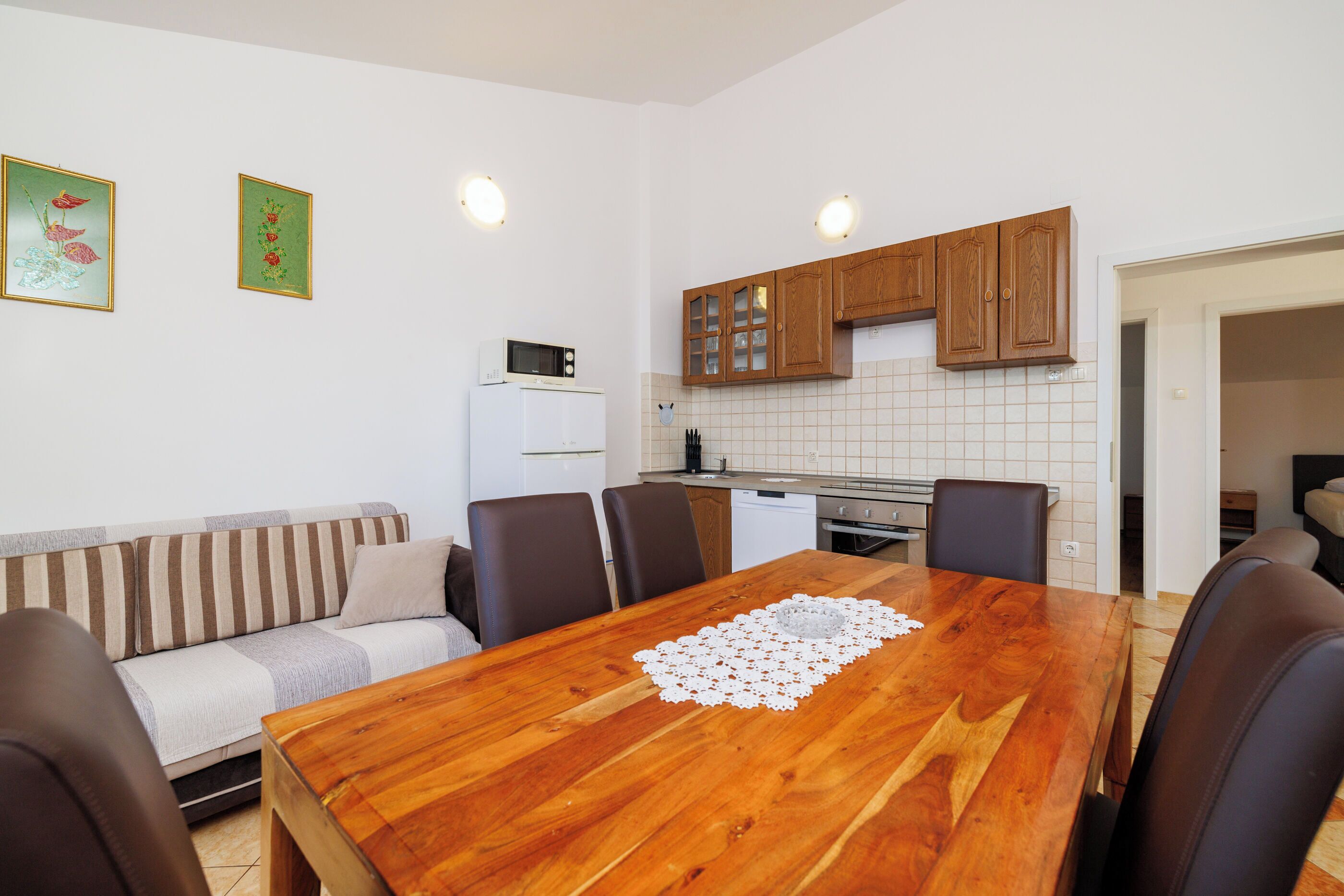 A dining area with a wooden table, chairs, a white refrigerator, and a kitchen in the background.