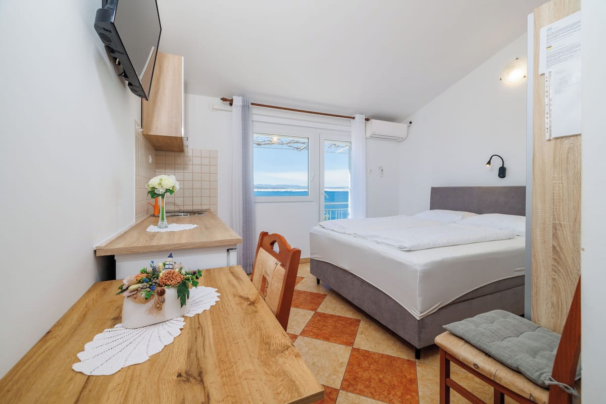 A bedroom with a bed, a wooden table with a vase of flowers, a chair, and a view of the sea through a window.