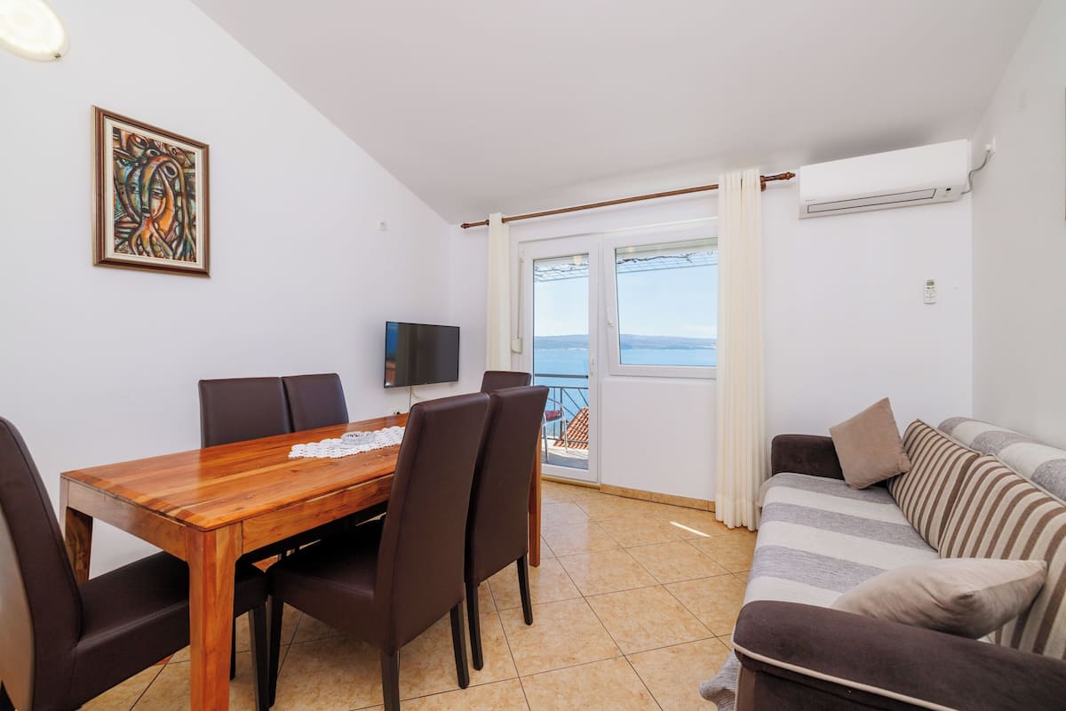 A dining area with a wooden table and chairs, a sofa, a TV, and a balcony with a view.