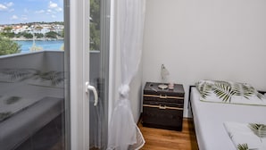 A balcony with a view of a coastal town, a bed with a leaf-patterned pillow, a bedside table with a lamp, and a small wooden chest. - Apartments Juro (Rogoznica)