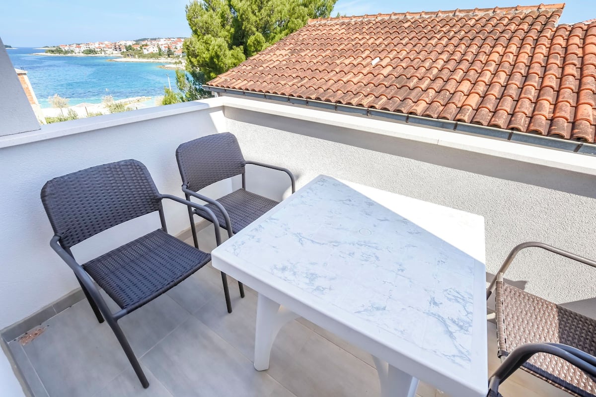 Un balcon doté d’une table en marbre blanc et de deux chaises noires, donnant sur une zone côtière avec un toit recouvert de tuiles.