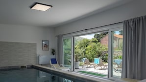 Una piscina interna moderna con pareti di vetro trasparenti, una luce a soffitto e vista su un giardino con lettini.