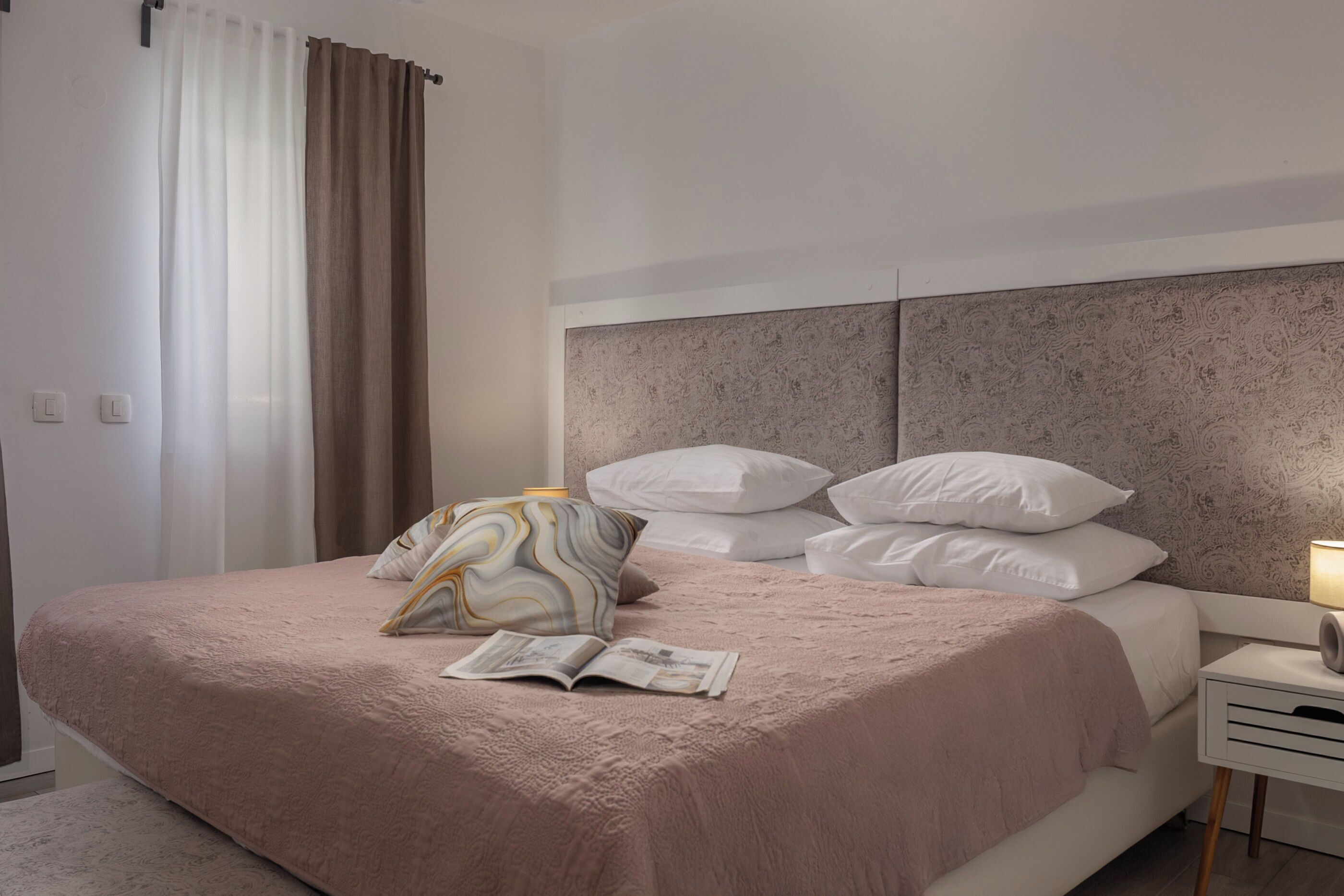 Un lit bien fait, recouvert d’une couvre-lit rose, avec des oreillers blancs et un coussin décoratif. On y trouve une table de chevet avec une lampe et un petit tiroir.
