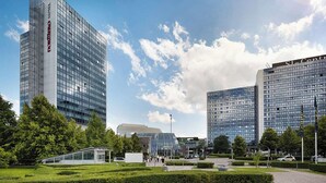 Et moderne hotel med en glasfacade, omgivet af velplejede grønne områder og andre moderne bygninger.