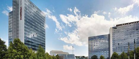 A modern hotel with a glass facade, surrounded by well-maintained greenery and other contemporary buildings.