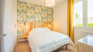 Une chambre à coucher avec un lit, une tête de lit en bois, une fenêtre avec des rideaux jaunes et un mur recouvert d’un papier peint à motifs de feuilles.