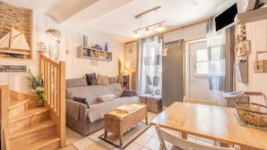 A cozy living room with a sofa, wooden coffee table, and a dining table. There is a staircase with wooden railings leading to a landing.