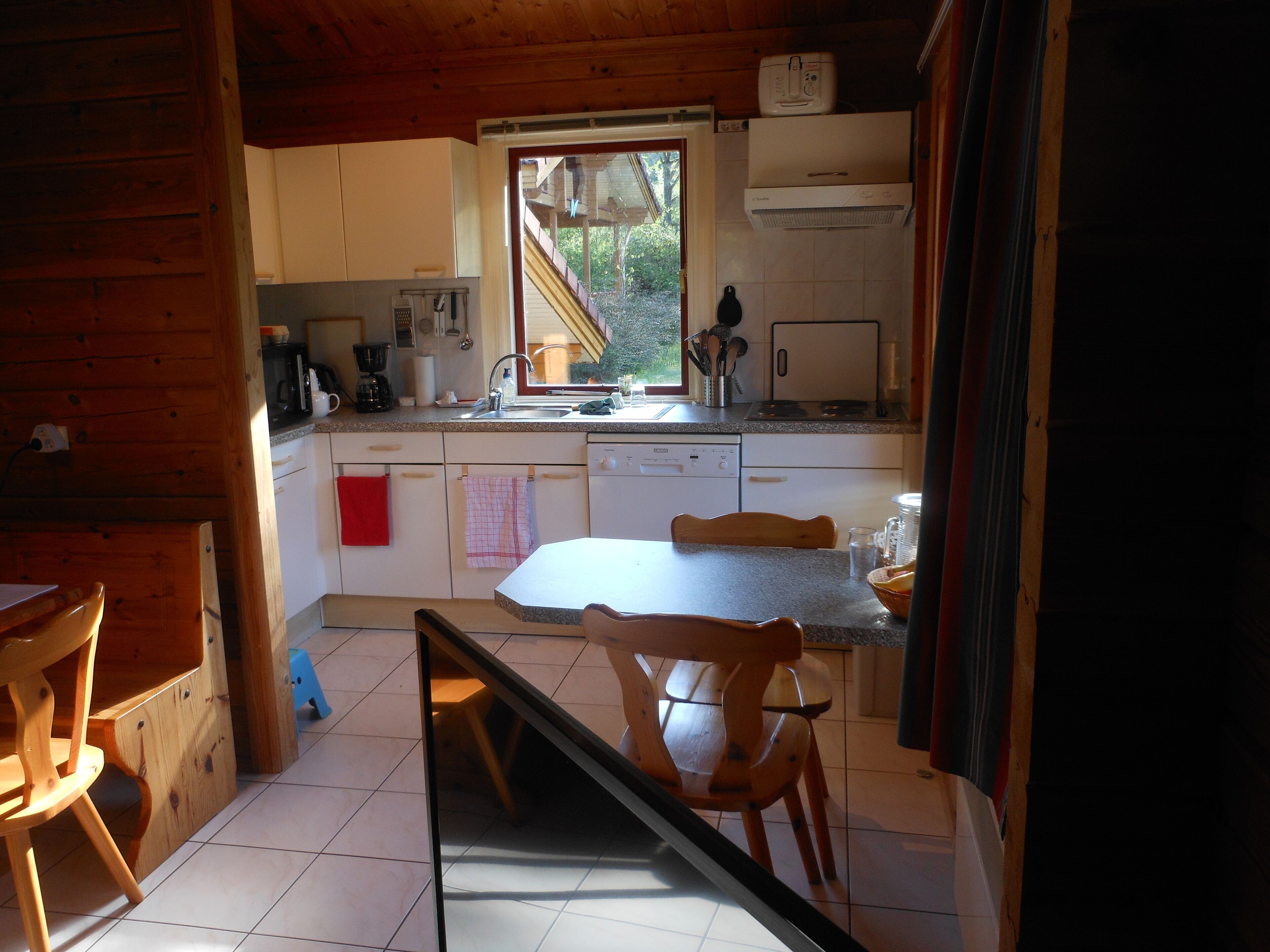 Een compacte keuken met houten kastjes, een wit aanrecht, een spoelbak, een vaatwasser en een raam met uitzicht op een tuin.