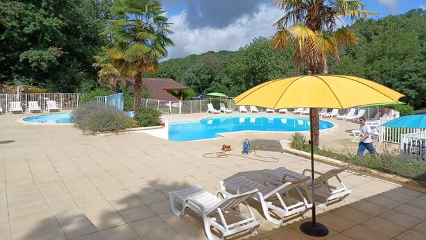 Un área de piscina con sillones de descanso y sombrillas, rodeada de árboles y un pequeño edificio.