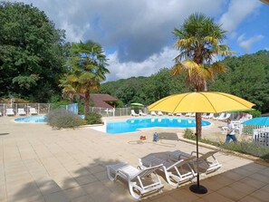 Un área de piscina con sillones de descanso y sombrillas, rodeada de árboles y un cielo despejado.