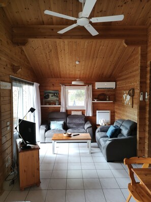 Interior acogedor de una cabaña con ventilador de techo, paredes de madera, un sofá gris, una mesa de centro de madera y un televisor.