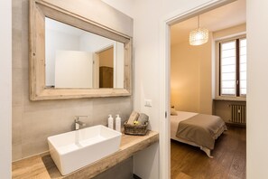 Un baño moderno con un espejo grande, un lavabo blanco y un mueble de tocador de madera. Se ve una cama a través de una puerta abierta al fondo.