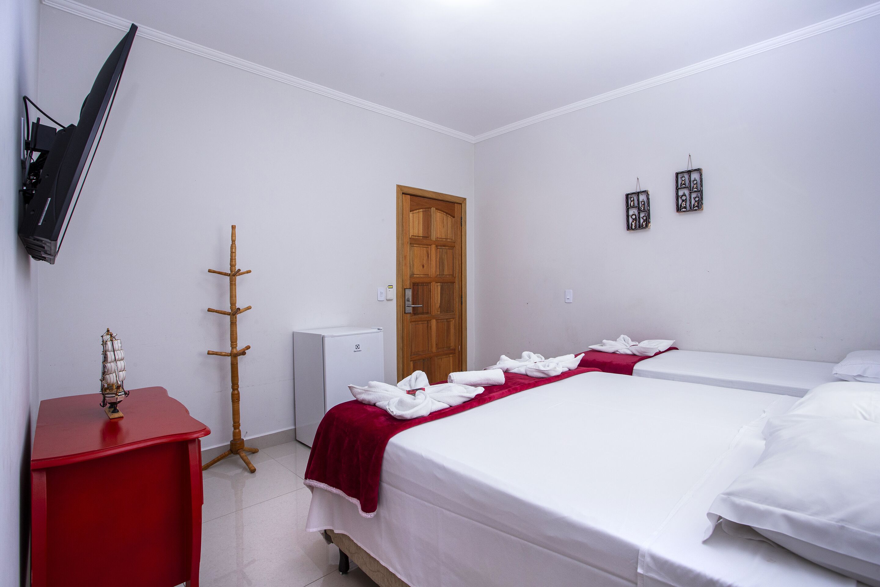 Quarto de hotel com cama, televisão presa na parede, cabideiro e pequeno frigorífico.