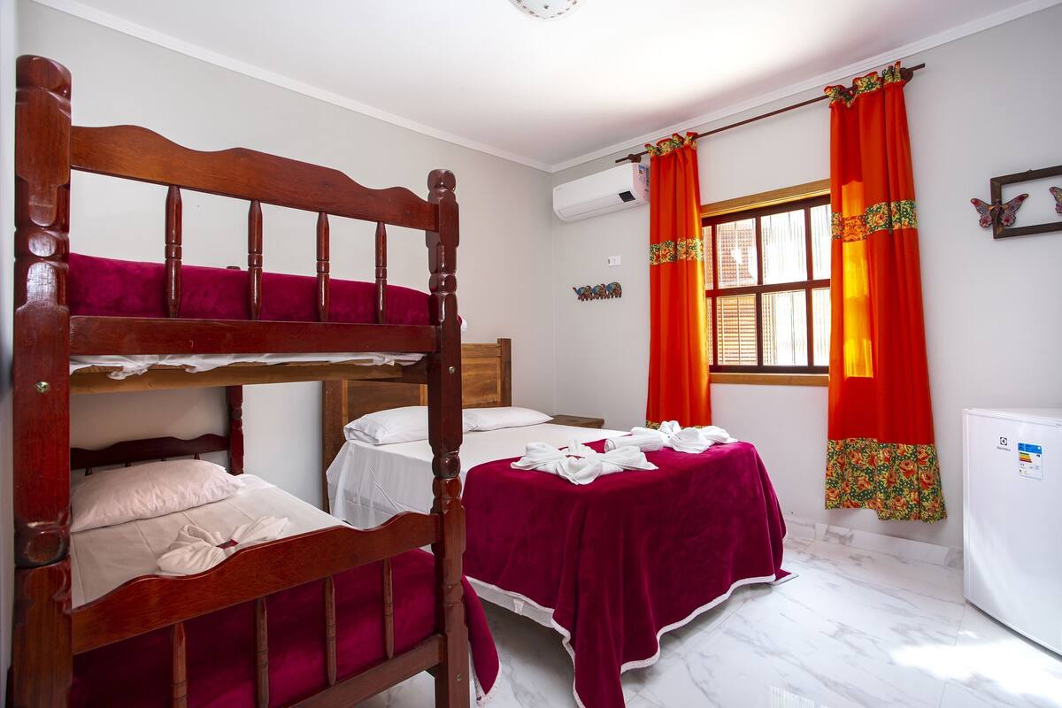 Una habitación con una cama litera, una cama individual, una colcha roja, una ventana con cortinas, un aire acondicionado instalado en la pared y un refrigerador.