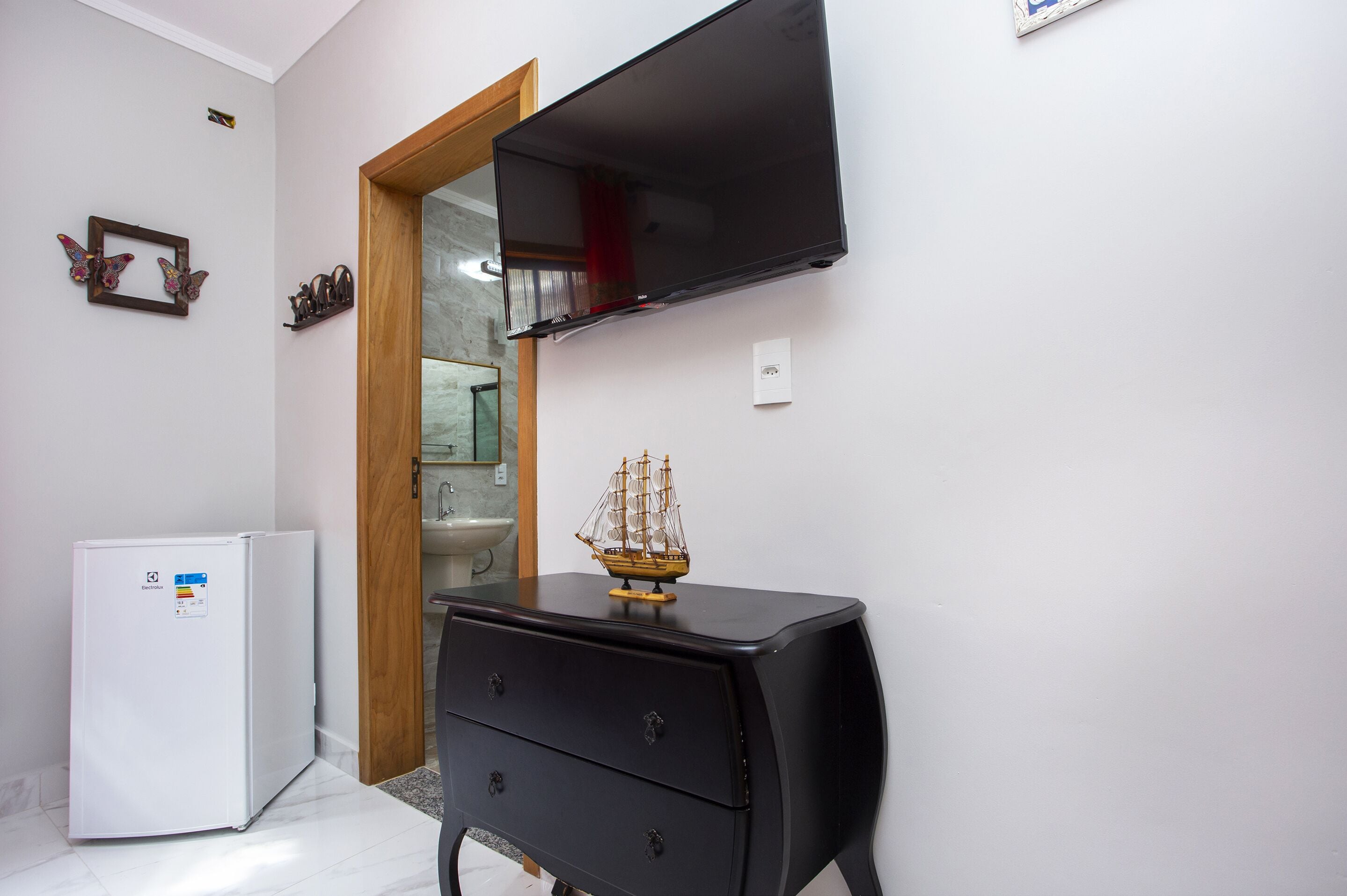 A compact room with a white refrigerator, a black dresser with drawers, a wooden door, a mounted flat-screen TV, and a small ship model on the dresser.
