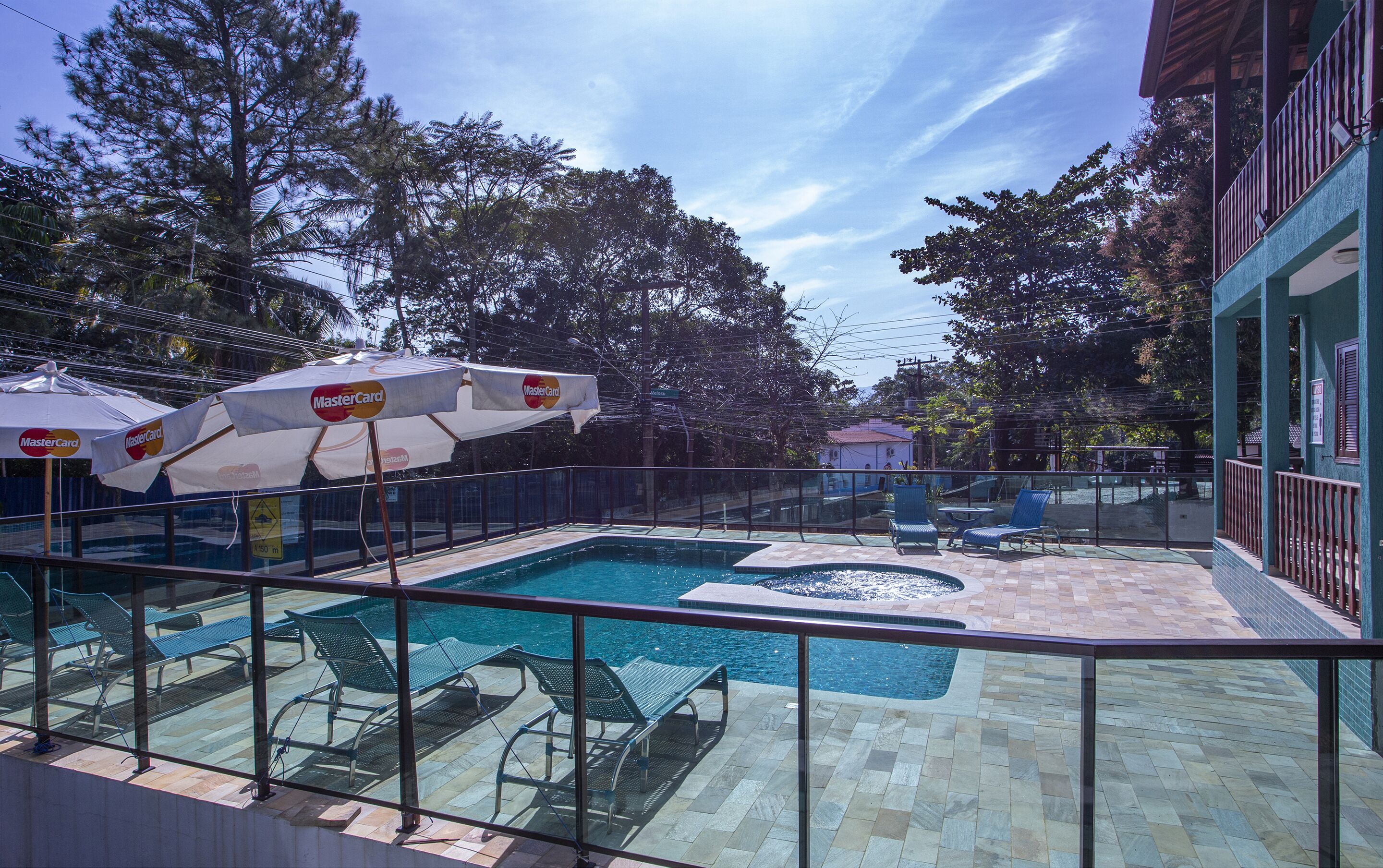 Un espace piscine aménagé avec des chaises longues et des parasols, entouré d’une clôture et d’arbres.