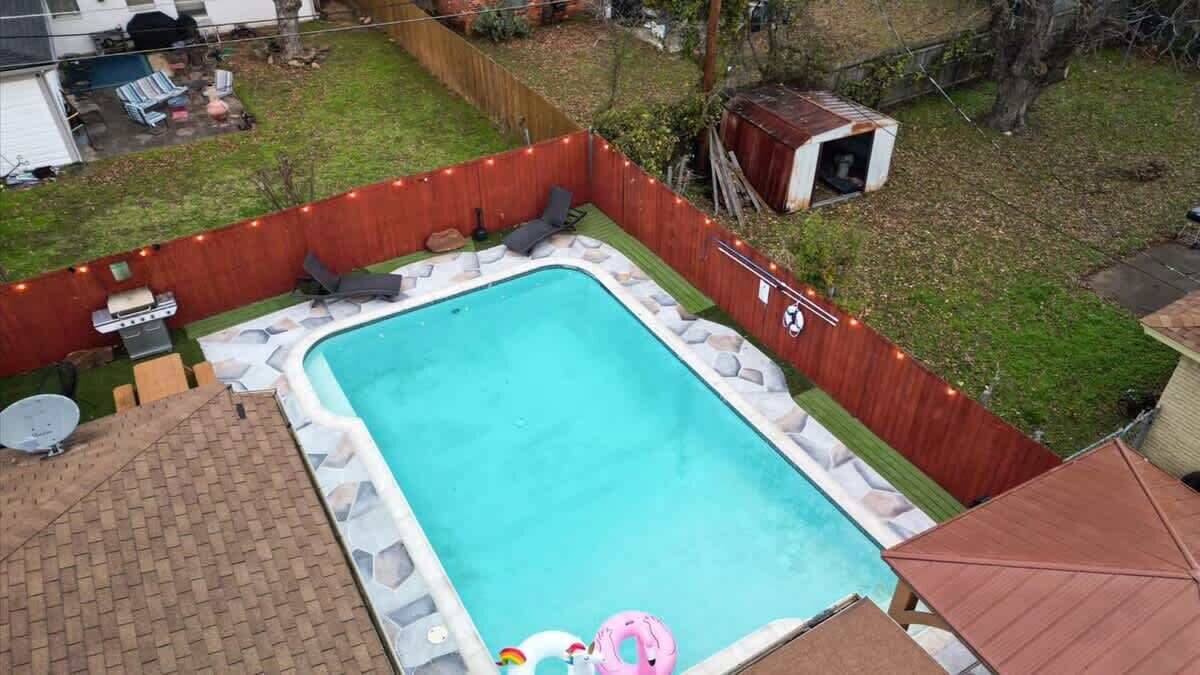A rectangular swimming pool with a wooden deck and lounge chairs, surrounded by a wooden fence and a small shed.