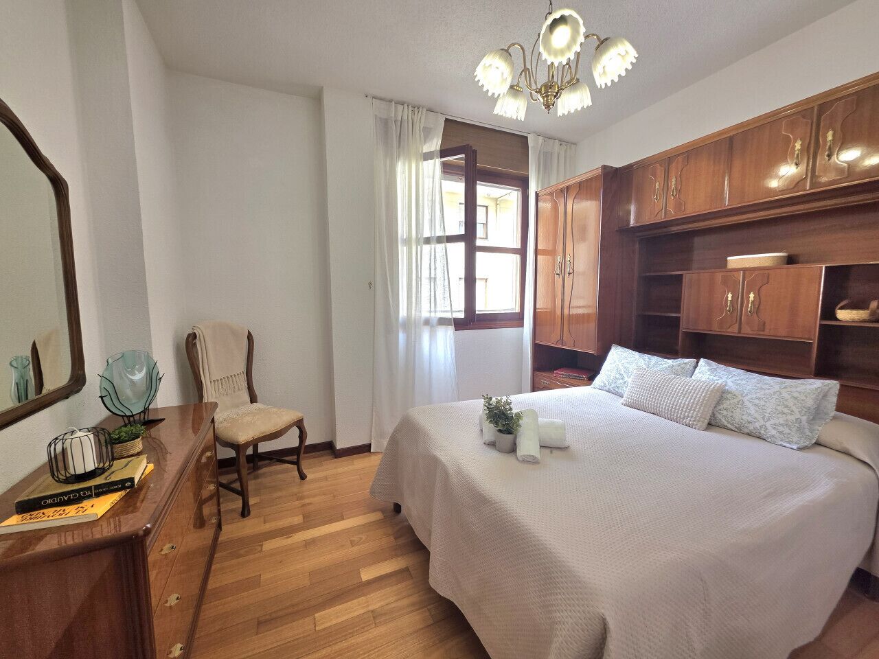 A bedroom with a bed, wooden wardrobe, a chair, a desk with books and a lamp, and a window with curtains.