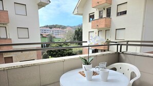 Una mesa redonda blanca con sillas en un balcón, una planta en maceta y vistas de edificios al fondo.