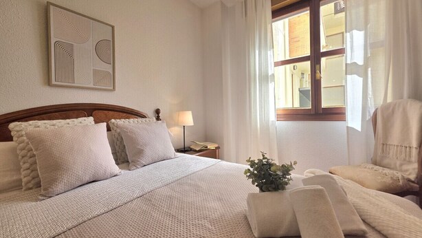 Habitación de hotel ordenada con cama, una silla, mesita de noche, lámpara y ventana con cortinas.