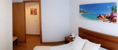 Quarto de hotel com uma cama, mesa de cabeceira, pintura de uma praia e uma porta de madeira.