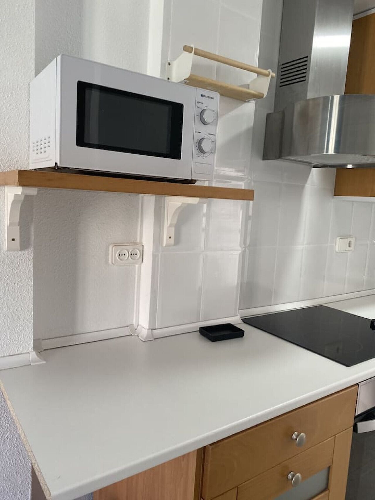 Une cuisine avec des armoires blanches, un four à micro-ondes posé sur une étagère en bois, un plan de travail noir et une hotte de cuisine en acier inoxydable.