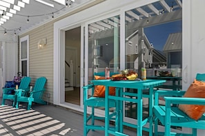 A covered outdoor area with turquoise chairs and a table, a glass door leading inside, and a view of a house in the background. - Beach+Pool+Hot Tub |  Golf Cart(incl) + Game Room (Destin)