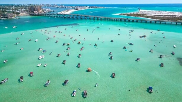 An aerial view of a coastal area with numerous boats on clear turquoise water, a pier extending into the sea, and a distant urban area with buildings. - Beach+Pool+Hot Tub |  Golf Cart(incl) + Game Room (Destin)