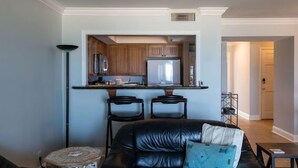 Uma sala de estar com um sofá de couro preto, uma mesinha redonda e uma área de cozinha com armários de madeira e uma geladeira.