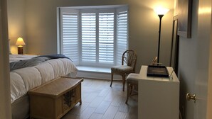Quarto com cama, mesa de cabeceira, cadeira, escrivaninha e janela com persianas.