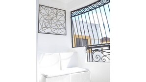 Een witte rotan stoel in een lichte kamer met een wanddecoratie met geometrische vormen en een verticaal verdeeld raam.