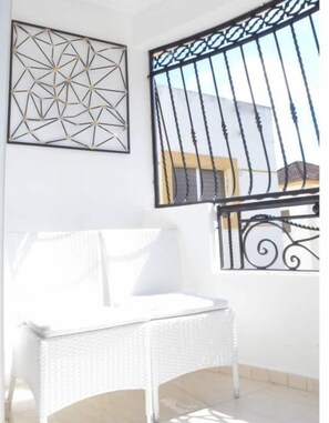Een witte rotan stoel in een lichte kamer met een wanddecoratie met geometrische vormen en een verticaal verdeeld raam.