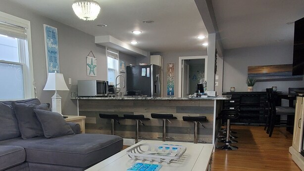 A modern living room with a grey sofa, a white coffee table with blue tiles, and a kitchen area with a bar counter and stools.