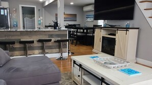 A modern living room with a grey sofa, a white coffee table, and a kitchen area with a TV and dining setup.