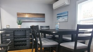 A dining area with a wooden floor, dark chairs with cushioned seats, a central table, and a wall-mounted air conditioner.
