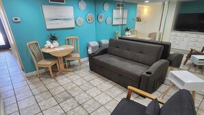 Una sala de estar con un sofá gris, sillas de madera, una mesita y suelo de baldosas.