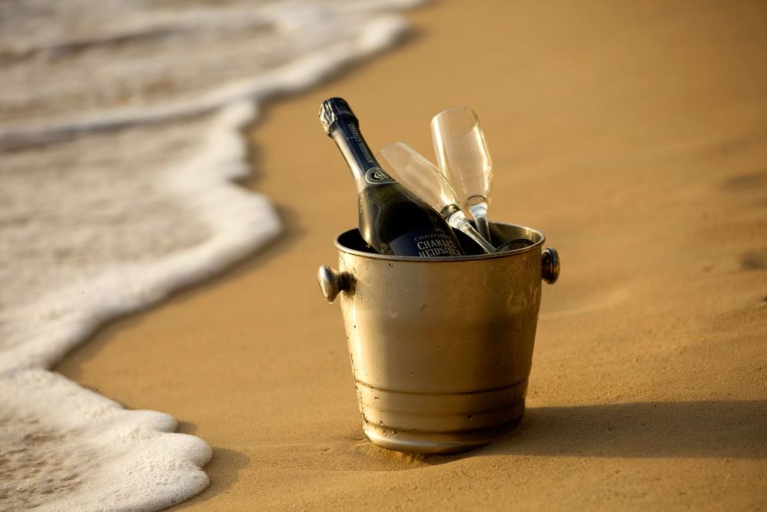 Un cubo metálico sobre una playa arenosa con una botella y vasos adentro.