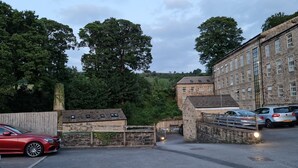 A parking area with a red car, stone buildings, and trees in the background. - 2 Bed Woodland Retreat - Garden & Free Parking (Keighley)