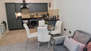 A compact kitchen with modern appliances, a dining area with a glass table, and a grey sofa with cushions. - 2 Bed Woodland Retreat - Garden & Free Parking (Keighley)