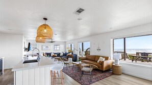 A spacious living area with a large window overlooking the ocean, a cozy seating area with a brown sofa and white chairs, and a kitchen with a white island and modern appliances. - Private Beach Walkover, Sunset Views & Room to Gather (Surfside Beach)