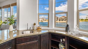 A kitchen with dark wood cabinets, a granite countertop, and large windows overlooking a body of water. - Lakefront Dream: Misters and The Best Corner Lot on the Lake! EV! (Indio)