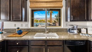 A kitchen with dark wood cabinets, a granite countertop, a window with a view of a pool and palm tree, and a coffee maker. - Lakefront Dream: Misters and The Best Corner Lot on the Lake! EV! (Indio)