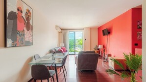 Un salon moderne avec une table à manger en verre, des canapés gris, un mur d’accent rouge, un téléviseur à écran plat et un balcon avec vue.