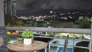 Un balcon avec une petite table et des chaises, un pot de fleurs et une vue sur le paysage urbain illuminé la nuit.