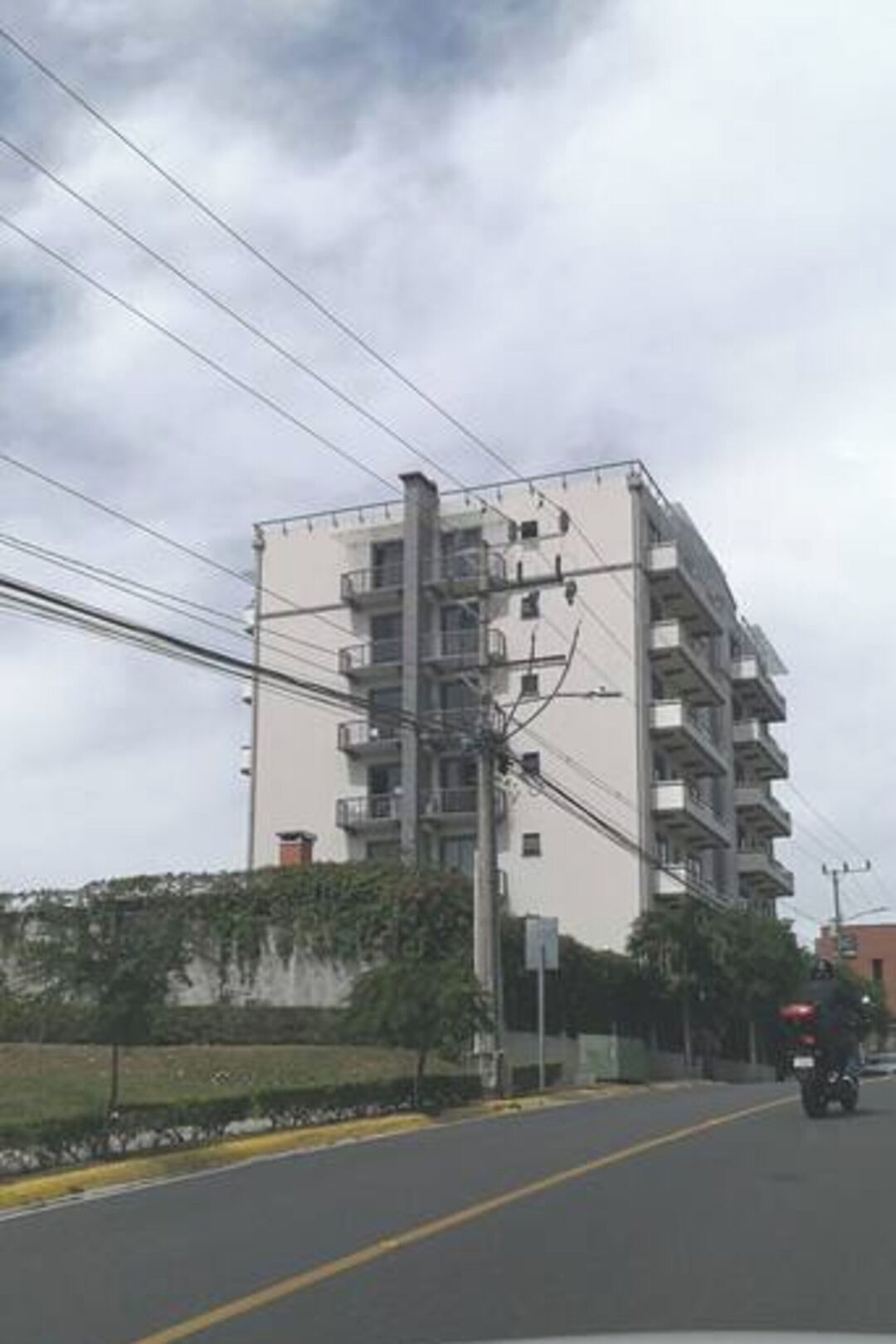 A multi-story building with balconies, a street in front, and a motorcyclist passing by.