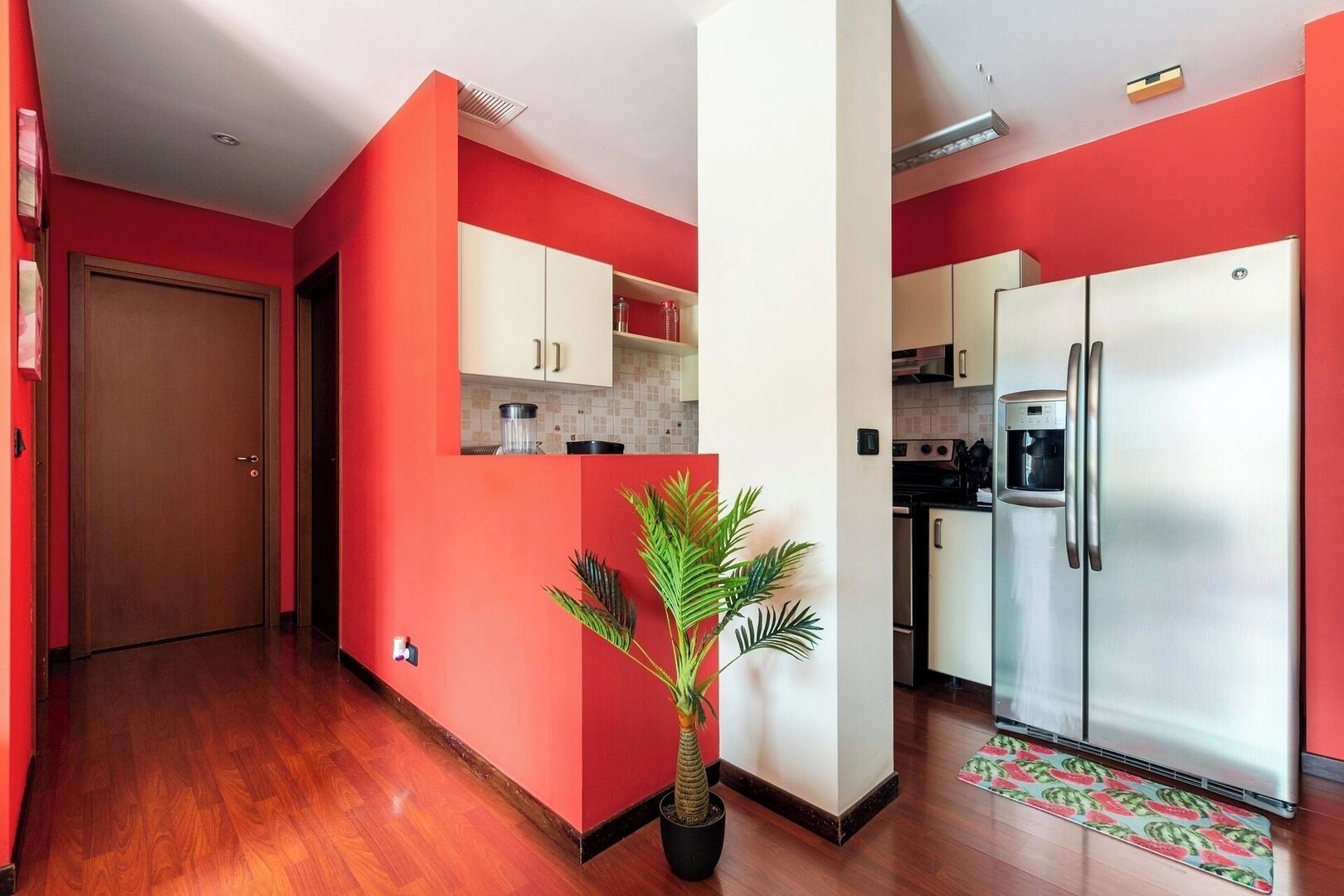 A compact kitchen with red walls, a wooden floor, a refrigerator, and a potted plant.