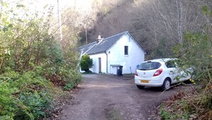 A rural road leading to a white house with a sloped roof, surrounded by dense vegetation.